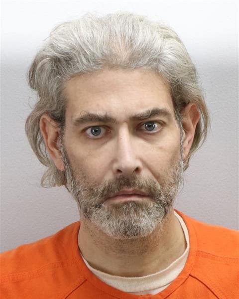 Mugshot of a white man with gray hair and a gray beard in an orange jumpsuit
